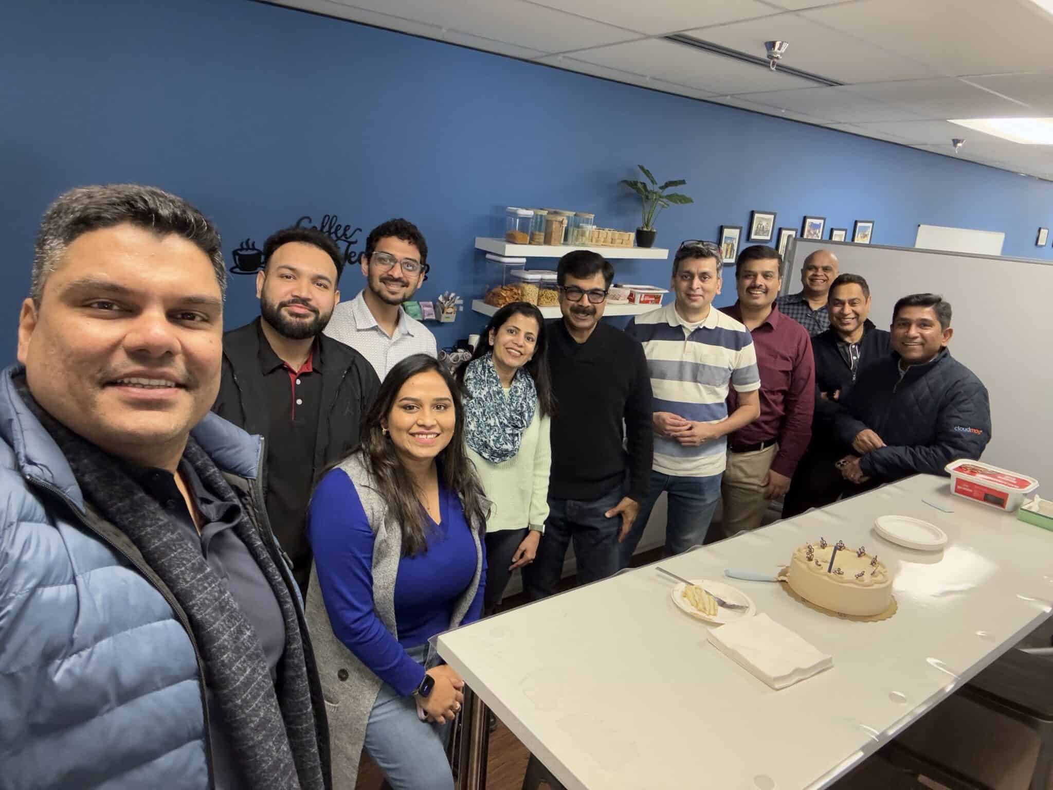 A group of people stand around a table with a cake and plates in an office break room, celebrating together and smiling at the camera—a great moment for any AI-First Consulting or Enterprise Intelligence team.
