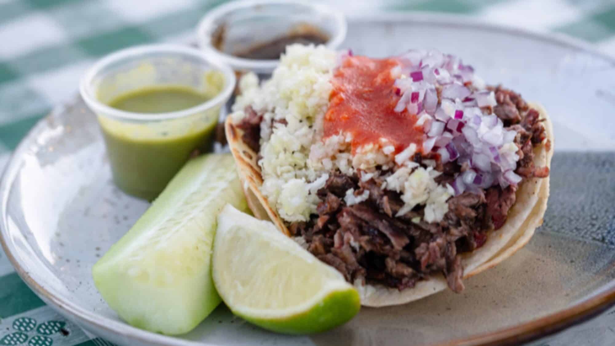 A plate with a taco filled with shredded meat, diced onions, cheese, and red salsa, served with cucumber, a lime wedge, and two plastic cups of sauce on the side.