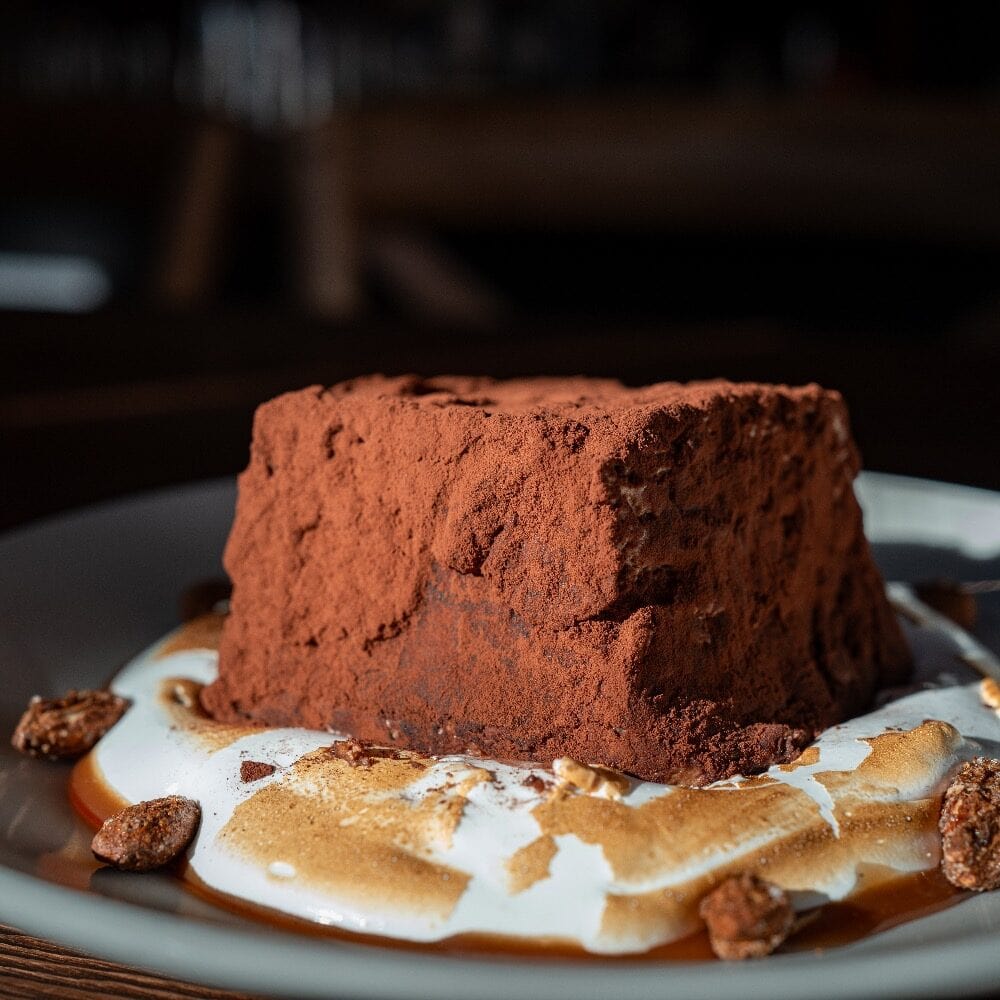 A chocolate mousse dessert dusted with cocoa powder sits on a plate with whipped cream, caramel sauce, and candied nuts.