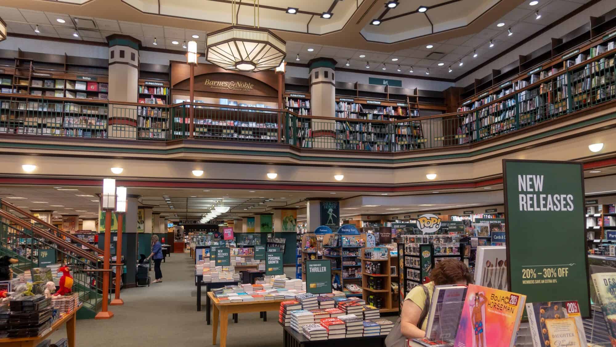 Interior of a two-story Barnes & Noble in downtown, featuring shelves of books, display tables, and a "New Releases" sign. A few people are browsing books and merchandise.