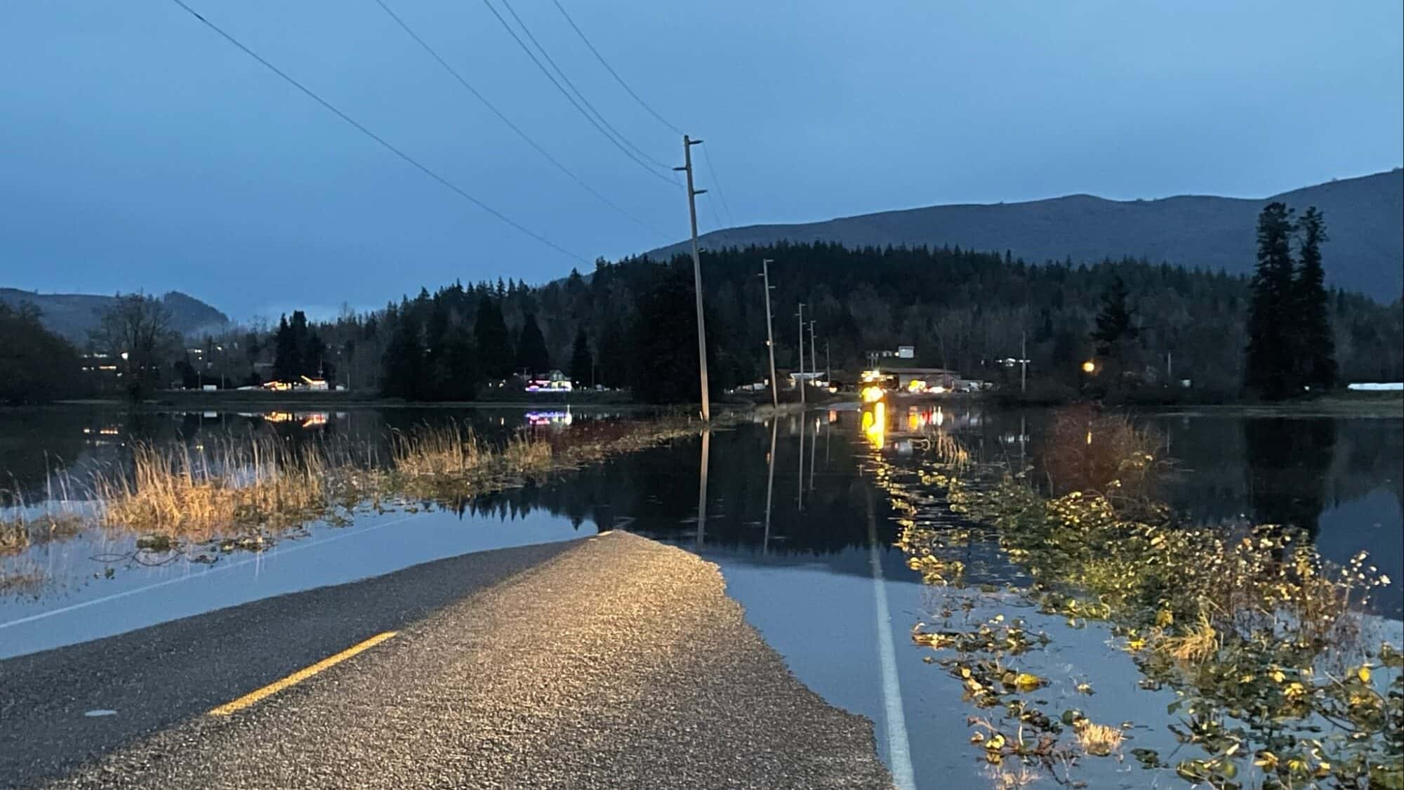 Flooded rural road at dusk with water covering both lanes, power lines overhead, and distant buildings reflecting lights on the water.