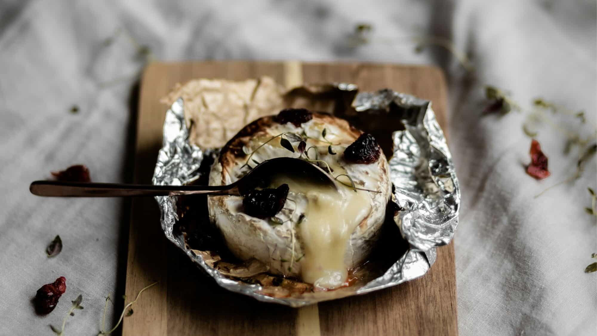 A baked round of soft cheese on a wooden board, partially wrapped in foil, with a spoon scooping out melted cheese. Dried herbs and berries are scattered on and around it.