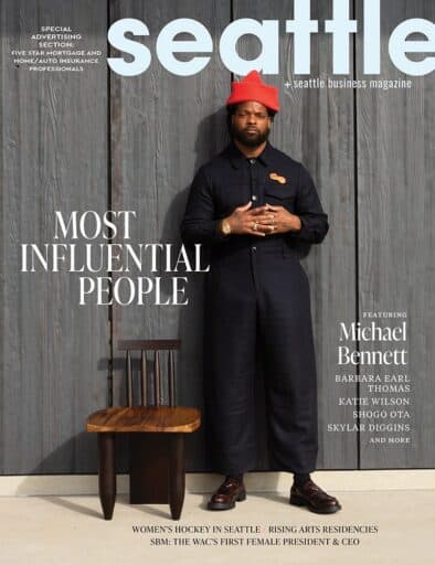 A man in a dark jumpsuit and red hat stands next to a wooden chair in front of a gray wall on the cover of Seattle Business Magazine's "Most Influential People" issue.
