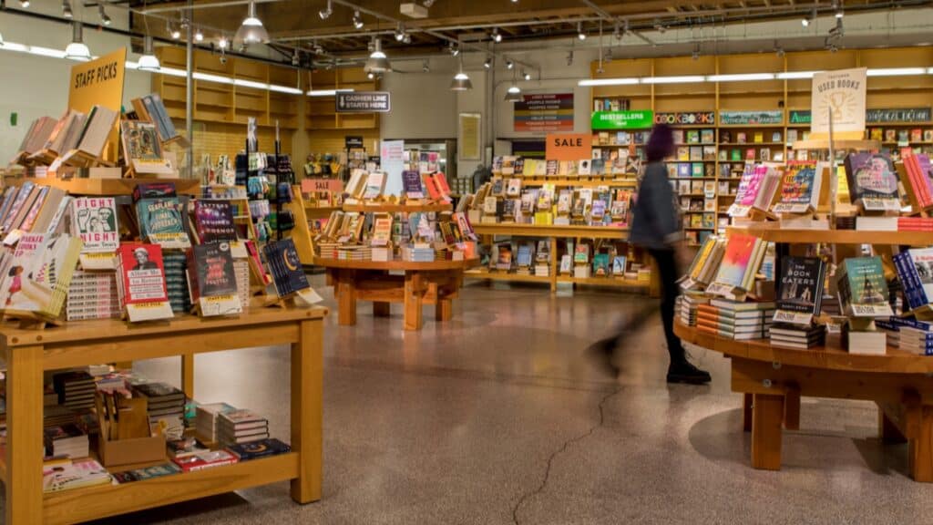 A spacious bookstore with wooden tables displaying various books, a SALE section in the background, and a person walking through the aisle.