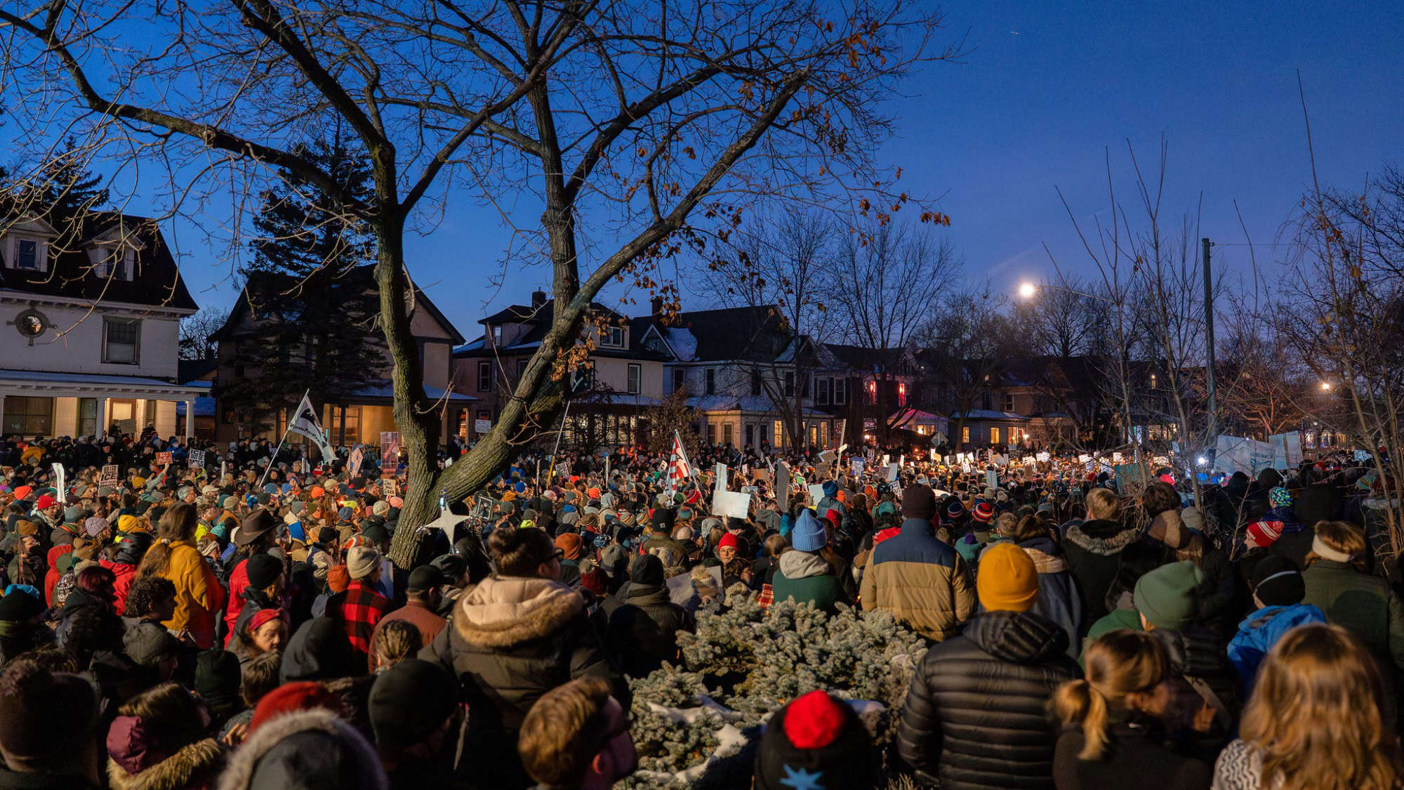 A large crowd gathers outdoors in a residential neighborhood at dusk, with people holding signs—perhaps seeking connection and coping with news overwhelm—while houses are visible in the background.