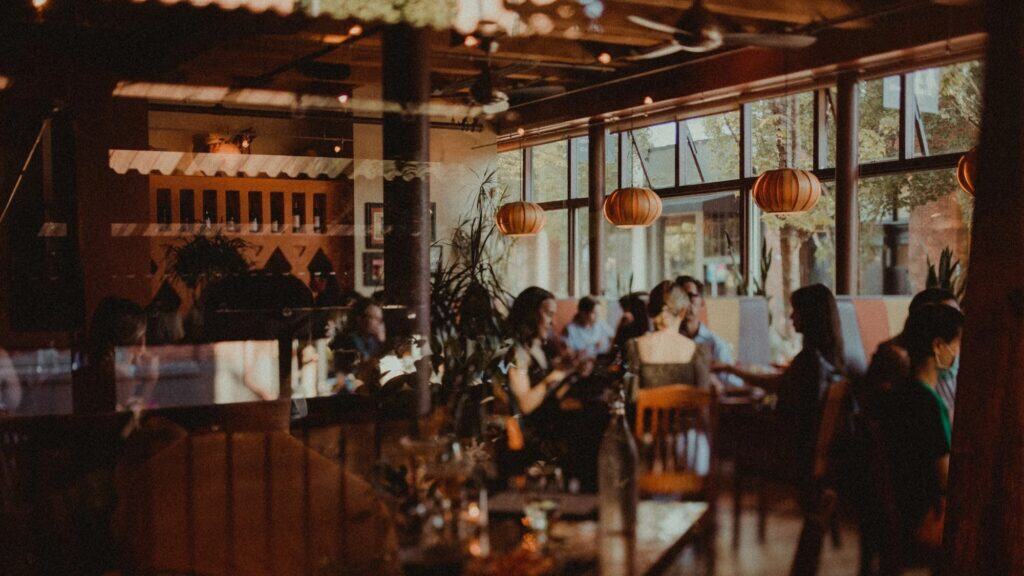 A group of people sit and converse at tables inside a warmly lit restaurant with large windows and hanging lantern-style lights.