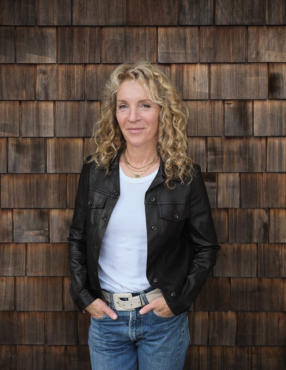 A woman with curly blonde hair stands in front of a wooden shingle wall, wearing a black jacket, white t-shirt, and blue jeans with hands in her pockets.