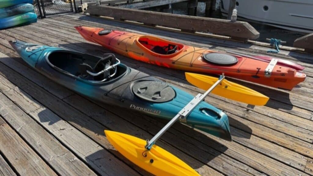 Two kayaks rest on a wooden dock; the front kayak is teal with yellow stabilizing outriggers, and the back kayak is orange.