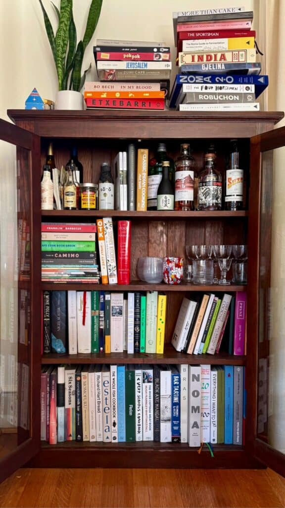A wooden cabinet with glass doors contains bottles and glasses on the middle shelf, and is filled with neatly arranged books on the top, middle, and bottom shelves. Stacks of books and a potted plant are on top.