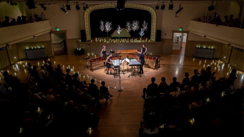 A marimba quartet performs on stage in a dimly lit concert hall, surrounded by an audience, with decorative lights and trees in the background.