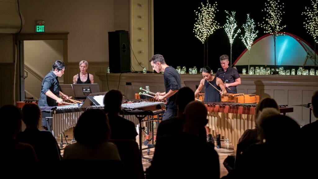 Four musicians perform on percussion instruments on a stage with illuminated decorations in the background, while an audience watches from seated rows.