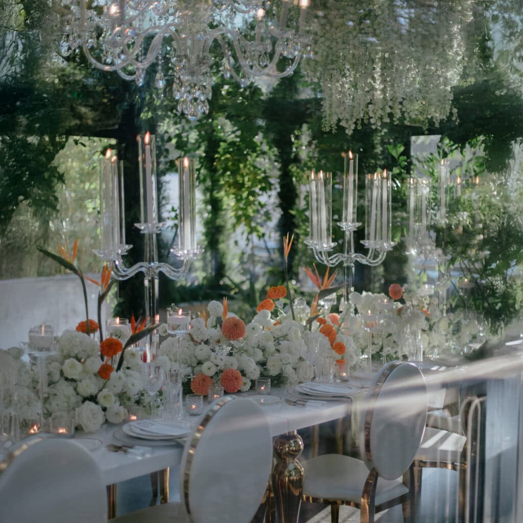 Elegant table setting with white and orange floral arrangements, tall crystal candelabras, white chairs, and a large chandelier, surrounded by greenery and reflective glass surfaces.