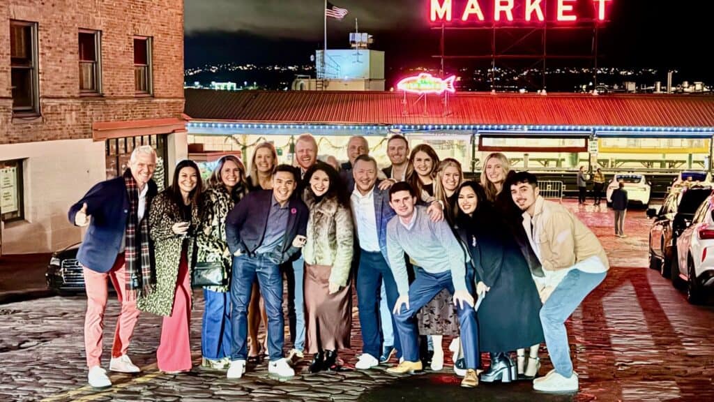 A group of people pose together on a cobblestone street at night in front of the illuminated Pike Place Market sign in Seattle.
