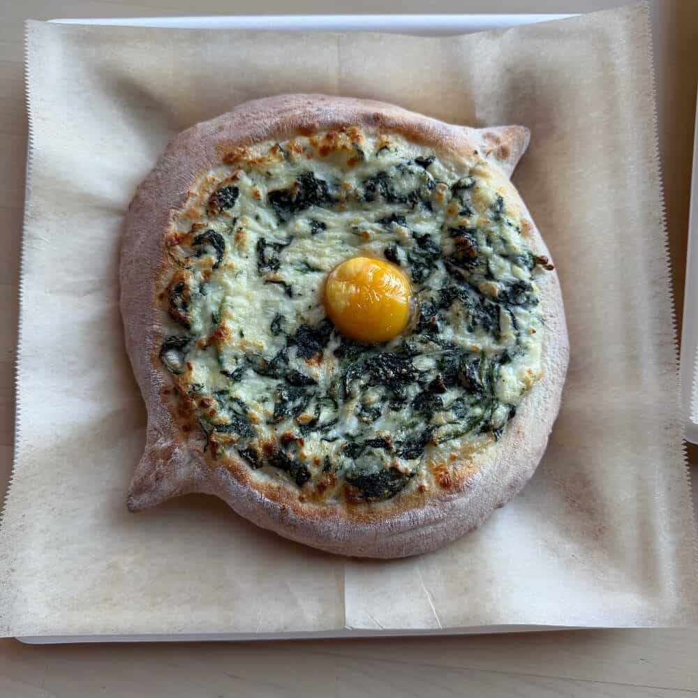 A round baked flatbread topped with melted cheese, cooked greens, and a whole egg yolk in the center, served on parchment paper.