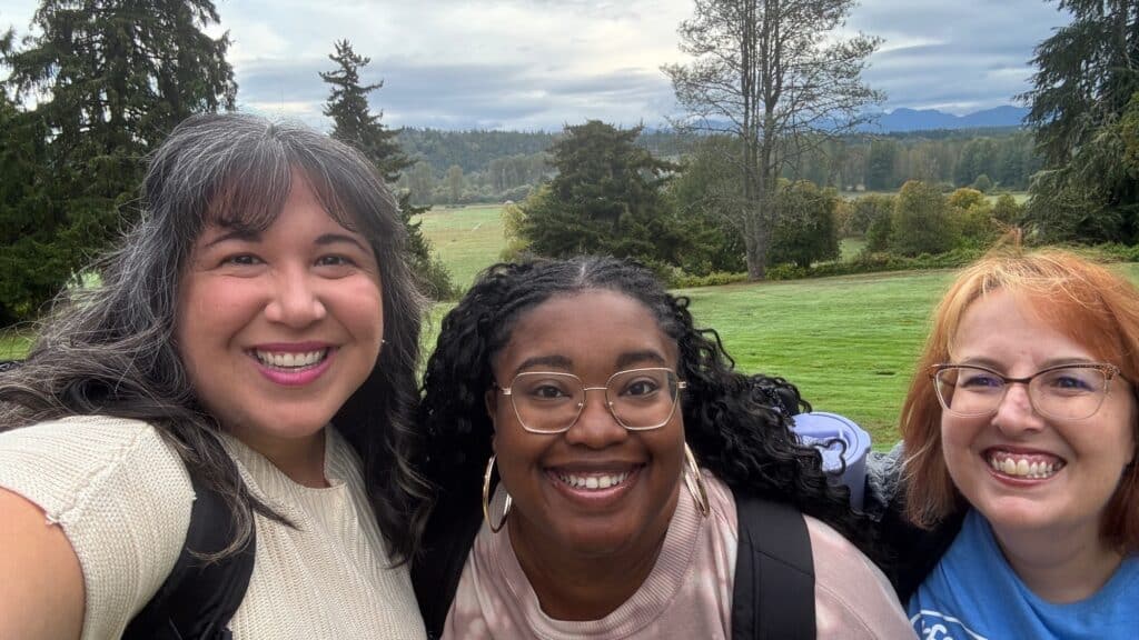 Three women smiling outdoors in a grassy area with trees and distant mountains in the background under a cloudy sky.