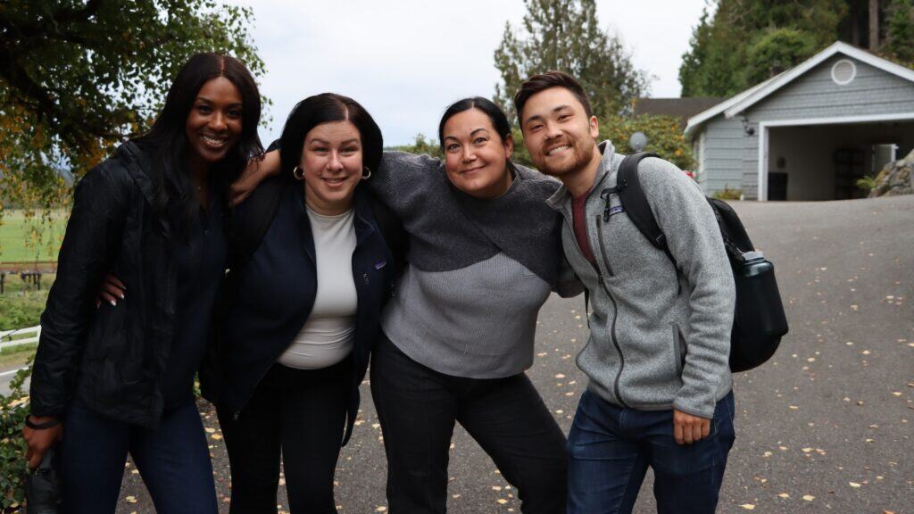 Four people stand closely together outdoors, smiling at the camera, with a gray house and trees in the background.
