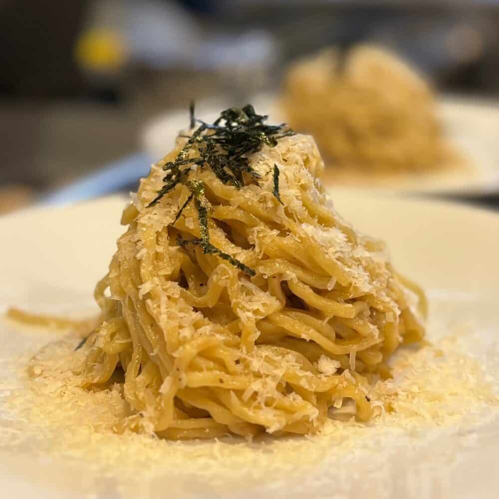 A close-up of a mound of pasta topped with grated cheese and thin strips of seaweed on a white plate.