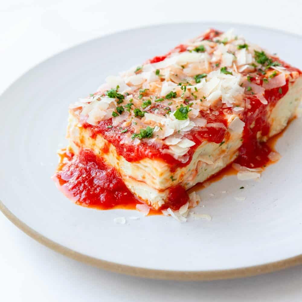 A slice of lasagna topped with tomato sauce, grated cheese, and chopped herbs is served on a white plate.