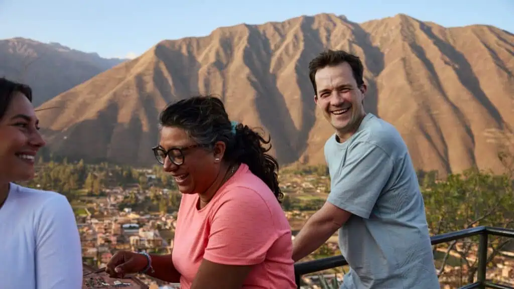 Three people stand outdoors on a balcony, laughing together with mountains and a town visible in the background.