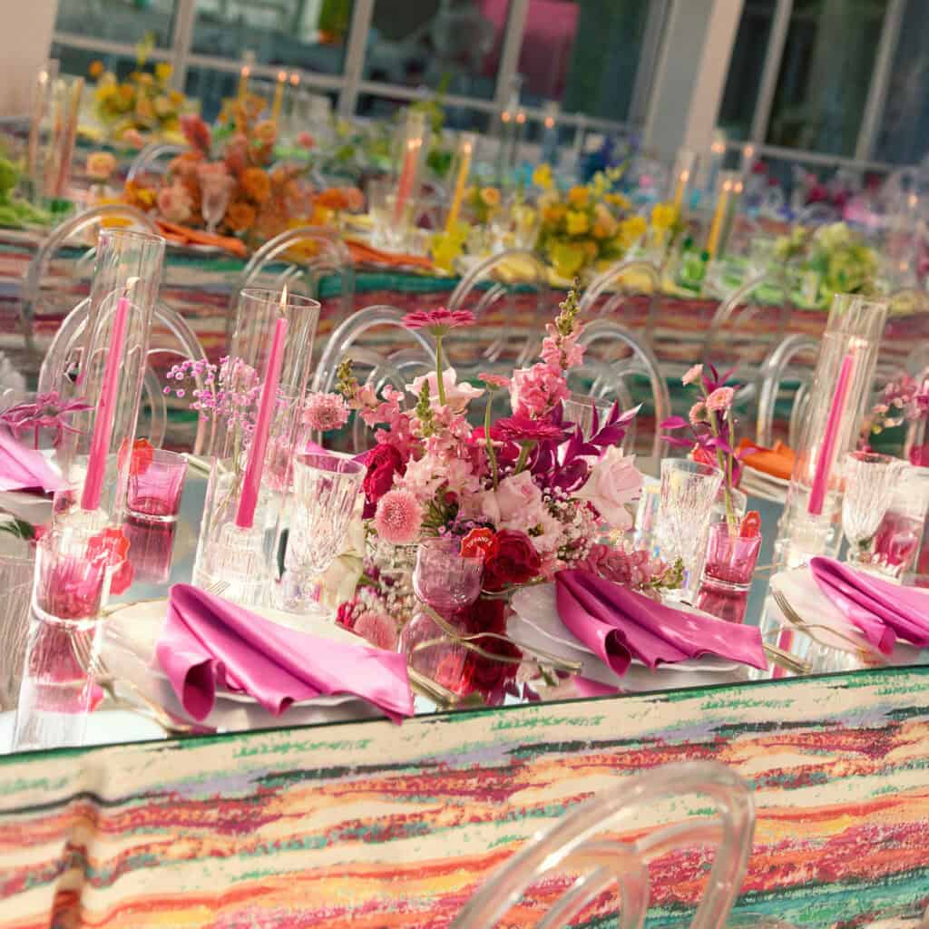 A long banquet table is set with colorful striped cloths, pink napkins, glassware, flowers, and candles in a bright, modern indoor space.