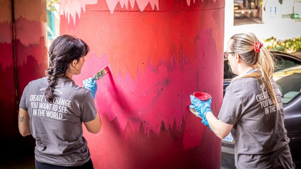 Two people wearing gloves and matching shirts paint a red mural on a large column outdoors.