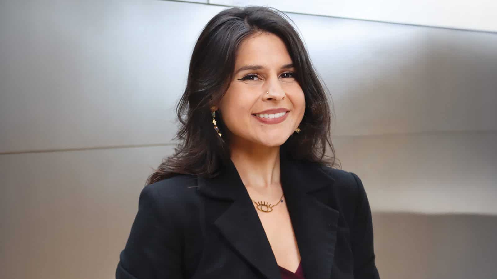 Councilmember Alexis Mercedes Rinck, a woman with long dark hair wearing a black blazer and gold necklace, smiles at the camera against a light metallic background.