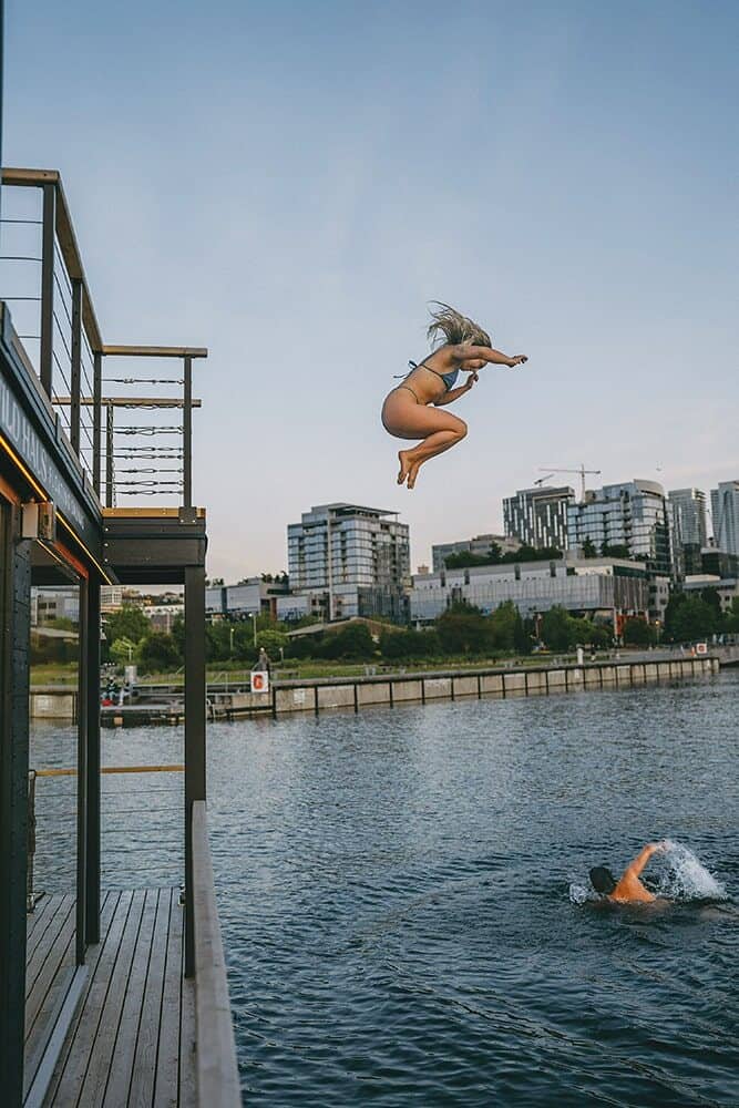 A person in a swimsuit jumps off a dock into a body of water while another swimmer is visible in the water below; city buildings are in the background.