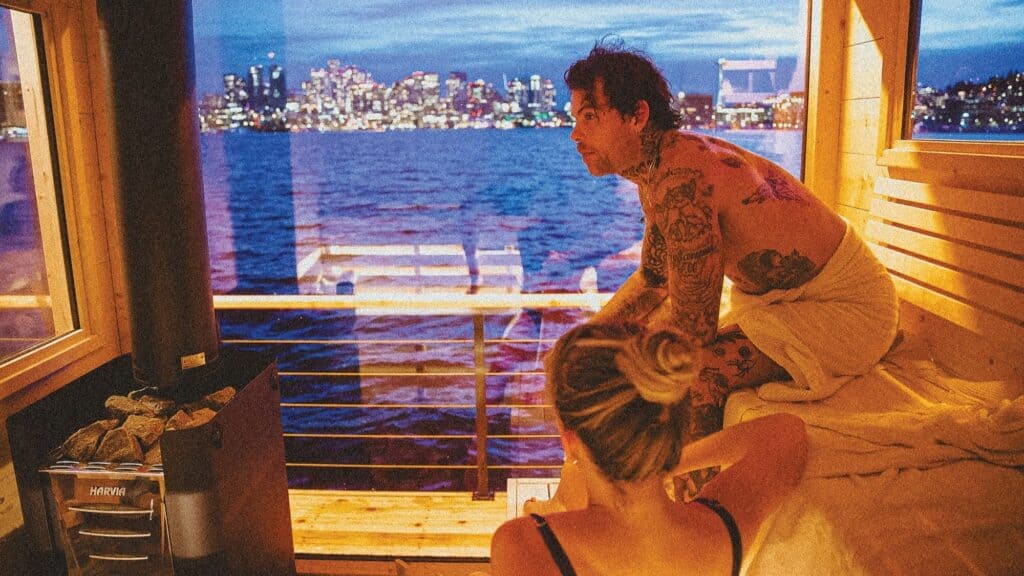 Two people relax inside a wooden sauna with large windows overlooking a body of water and a city skyline at dusk.