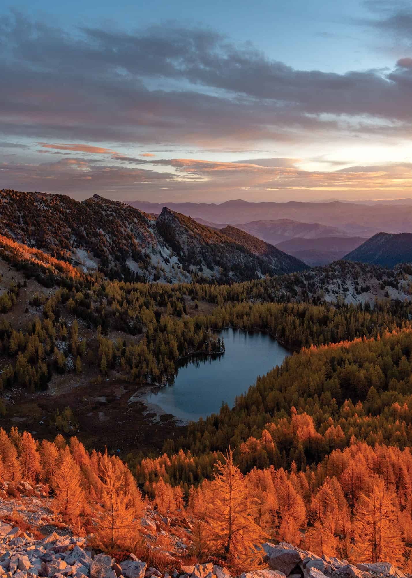 A mountain landscape at sunset shows a small lake surrounded by autumn trees and distant ridges under a partly cloudy sky, perfect for hiking or photography with Tiffanie Yang.