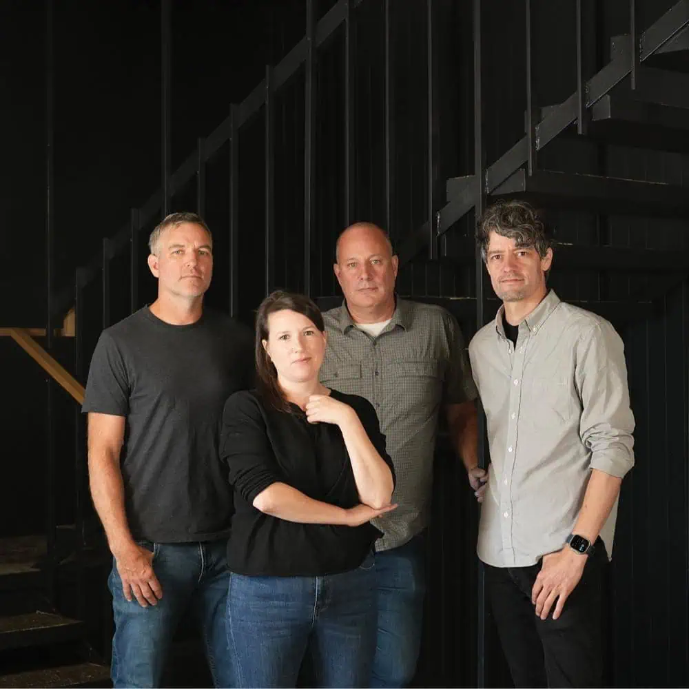 Four adults stand together indoors in front of a dark staircase, posing for a group photo—a clear display of joint effort and camaraderie at SBM.
