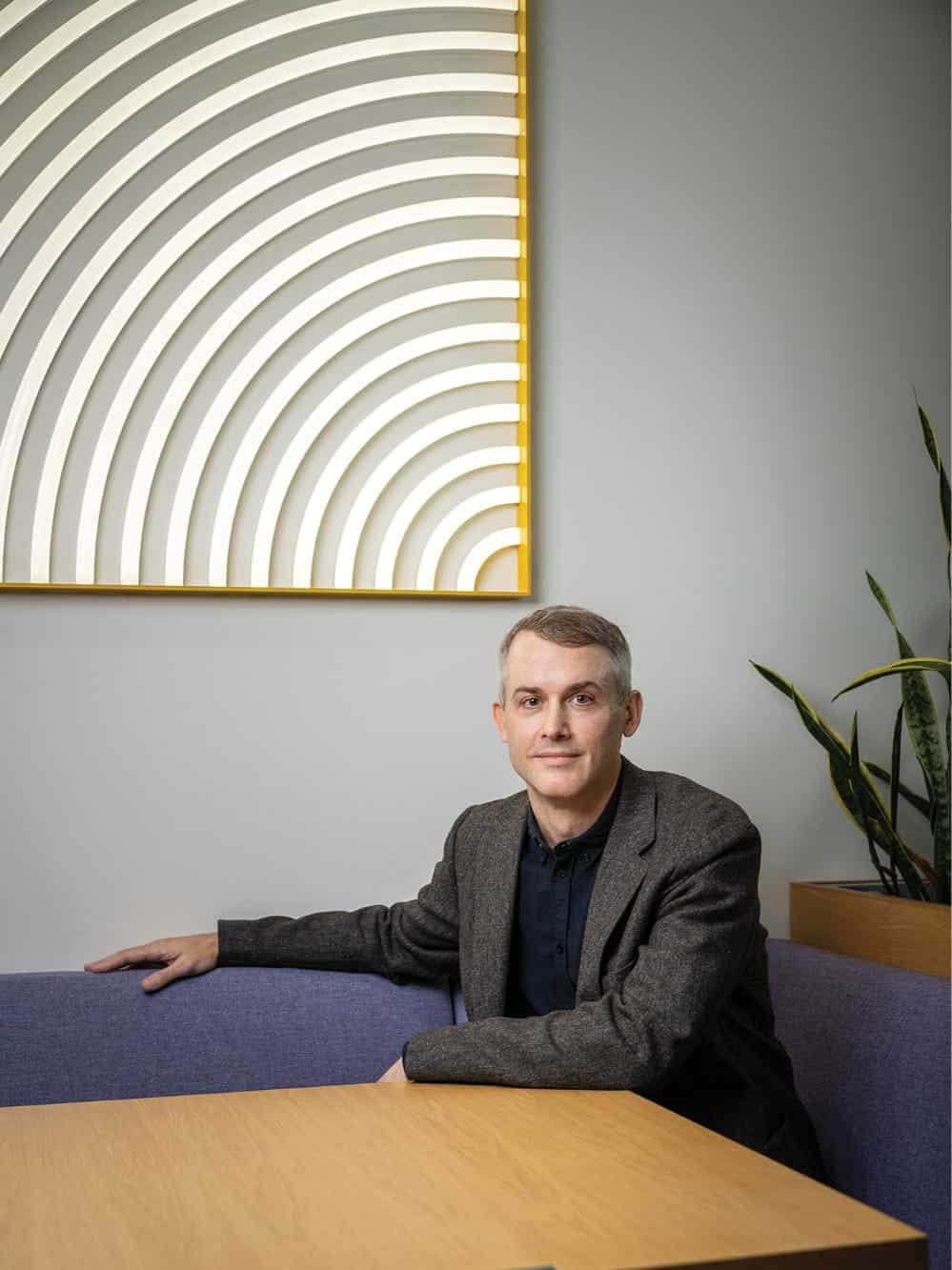 A man in a gray blazer sits at a wooden table with a blue bench, next to a plant, in front of the Eye Eye optometry clinic’s wall art—a geometric piece with white and gold curved lines, reflecting Dr. Will Pentecost's modern vision.