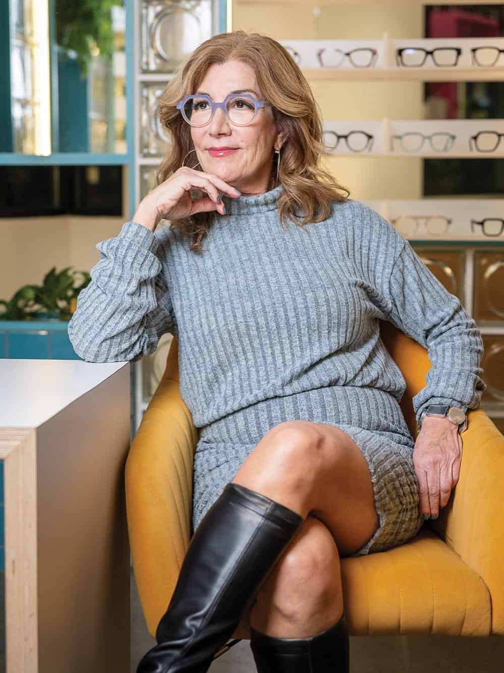 A woman with wavy, light brown hair and blue glasses sits in a yellow chair at optometry clinic Eye Eye, wearing a gray knit dress and black boots. Eyeglass frames are displayed in the background, curated by Dr. Will Pentecost.
