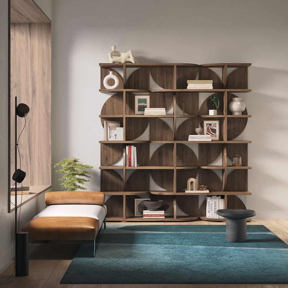 Modern living space with a geometric wooden bookshelf, books, and decor items—a stylish joint effort of design elements—plus a tan and white chaise lounge, side table, floor lamp, and blue area rug.