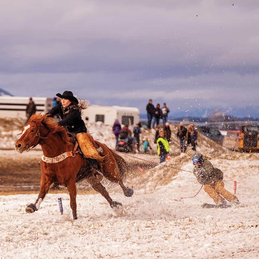 A person rides a horse at speed across snow while another person on skis is pulled behind, with spectators and vehicles in the background.