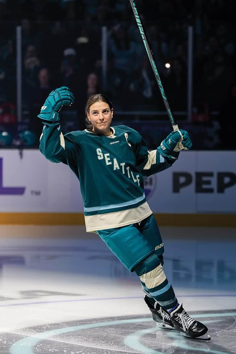 A hockey player in a teal and white Seattle jersey celebrates on the ice, raising one arm and holding a hockey stick.