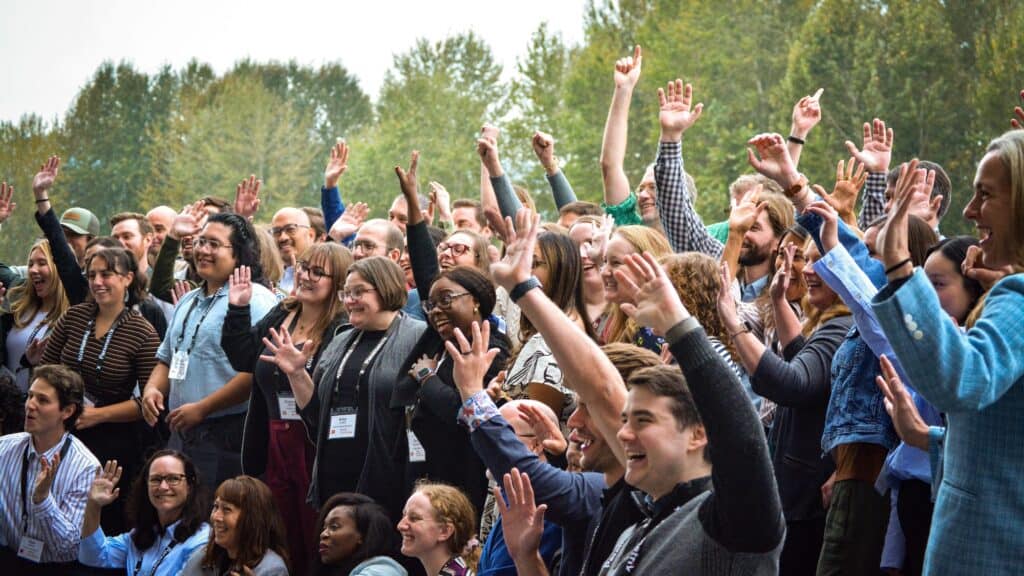 A large group of people standing outdoors with trees in the background, many of them smiling and raising their hands, wearing name badges.