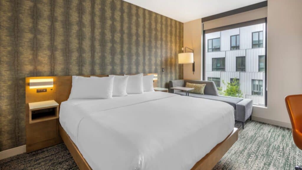 Modern hotel room with a large bed, white bedding, gray sofa, and urban views—perfect for travelers exploring Portland's Pearl District. Neutral-toned decor and cozy touches make it an inviting retreat featured in the Wanderer’s Guide.