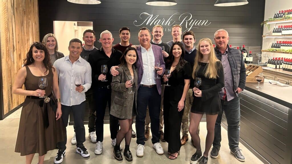A group of people pose together indoors, holding wine glasses, in front of a wall with the sign "Mark Ryan" and shelves of wine bottles.