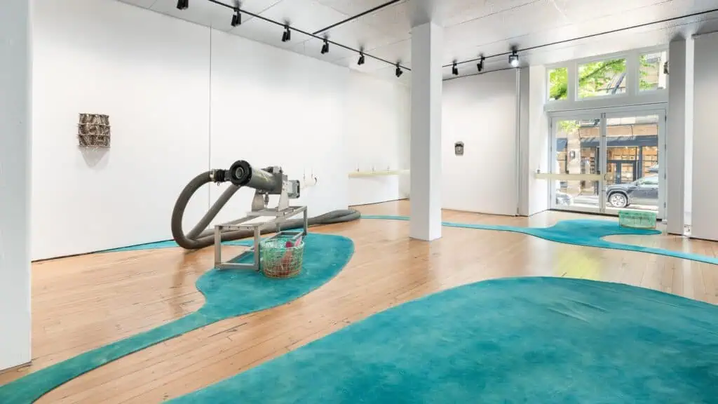 Art gallery in Portland’s Pearl District with white walls, wooden floors, blue-green carpet accents, and an industrial sculpture with tubing; artwork lines the walls and street views beckon through large windows—a true Wanderer’s Guide gem.