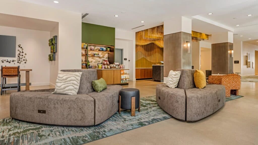 Modern hotel lobby in Portland’s Pearl District with two curved gray couches, decorative pillows, a small stool, snack bar in the background, and warm lighting—perfect for any travel guide.