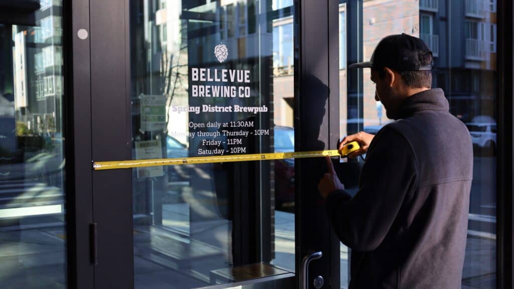A man measures the glass door of Bellevue Brewing Co. Spring District Brewpub with a tape measure; business hours are displayed on the door.