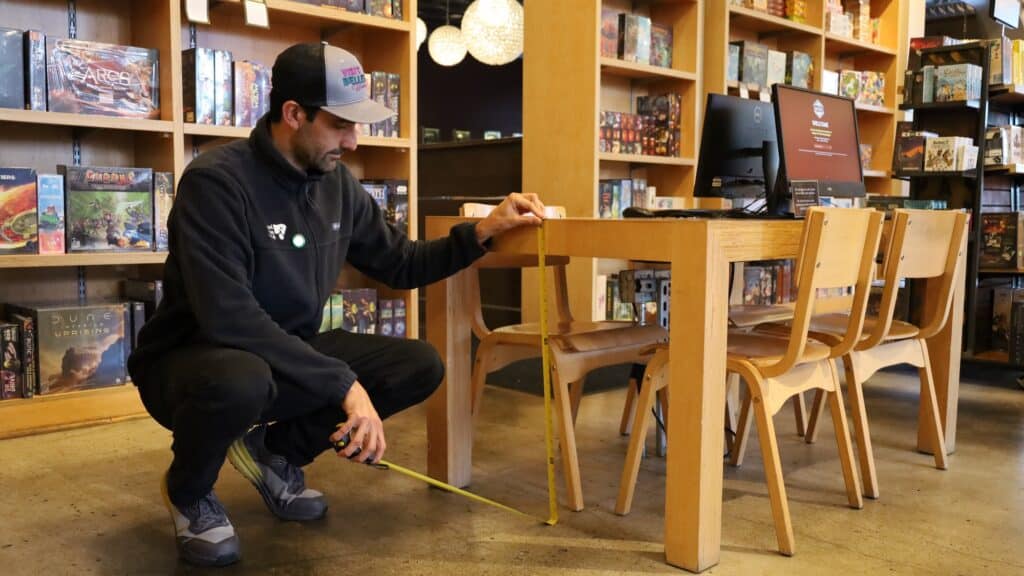 A person measures the height of a wooden table with a measuring tape in a game store, surrounded by shelves filled with board games.