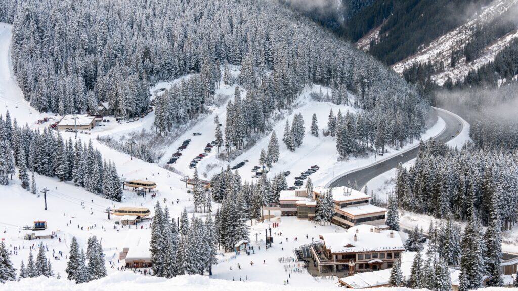 A snow-covered ski resort with multiple buildings, a winding road, parked cars, and people skiing among pine trees on a mountain slope.