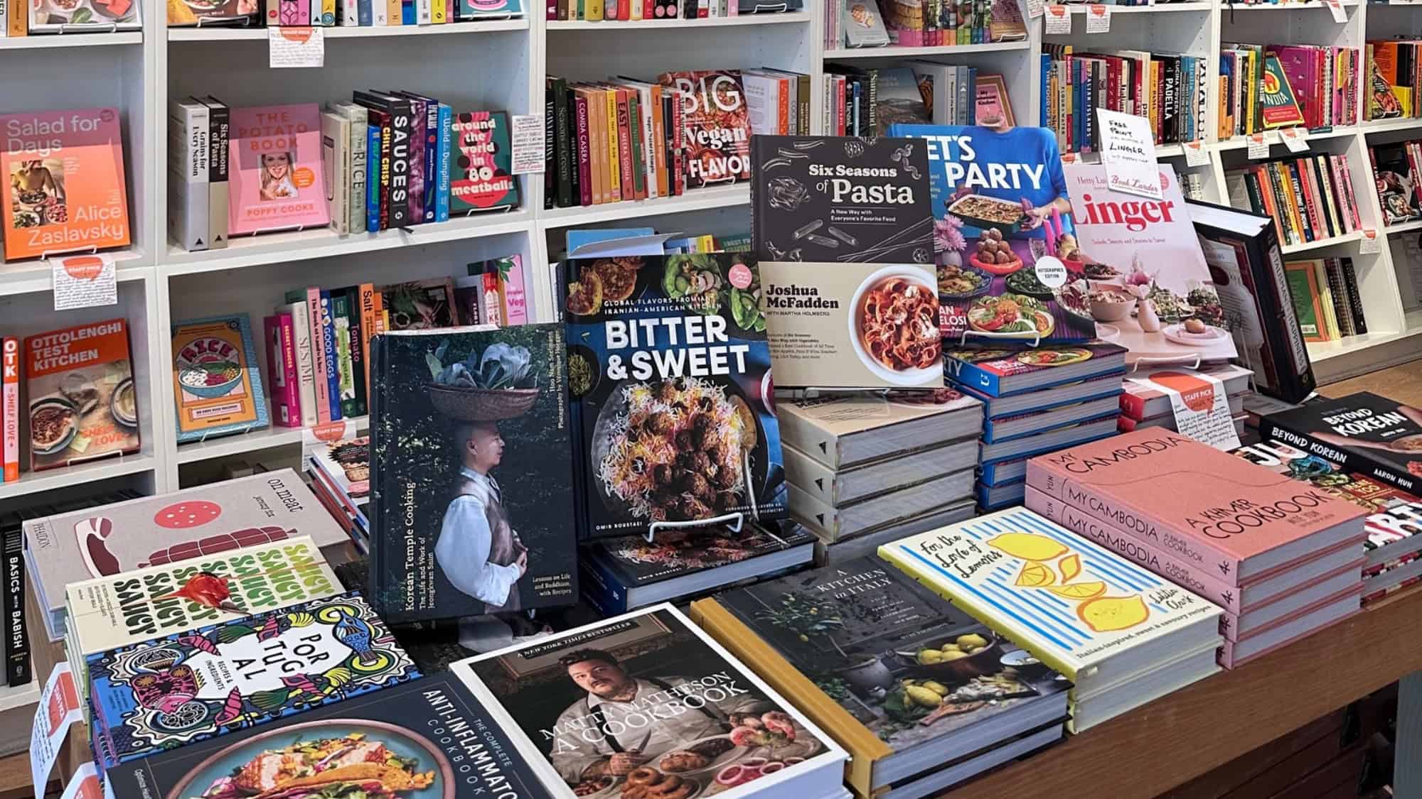 A display table with an enticing cookbook selection, including titles like "Bitter & Sweet," "Six Seasons: Pasta," and "Pet’s Party," perfect for date night inspiration, in front of shelves filled with more books.