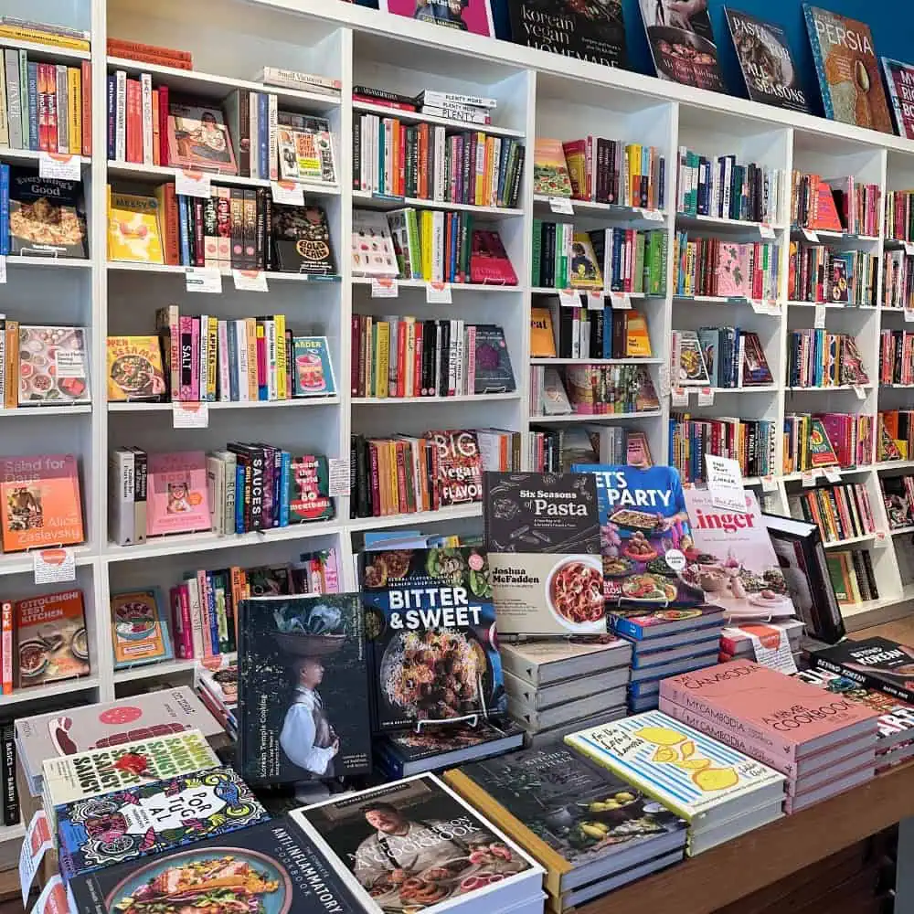 Bookshelves and a table display a variety of colorful cookbooks and food-related titles, perfect for planning your next Date Night or Restaurant Roundup, all in a well-lit bookstore.