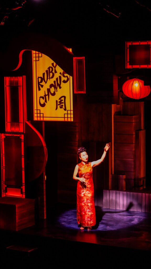 A performer in a red dress stands on stage under dramatic red lighting, with a "Ruby Chou's" sign and lanterns in the background.