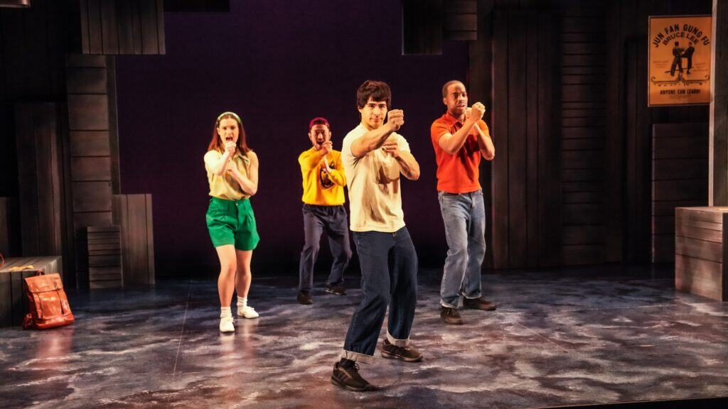 Four people stand on a stage in mid-action poses, facing forward, with dramatic lighting and wooden set pieces in the background.