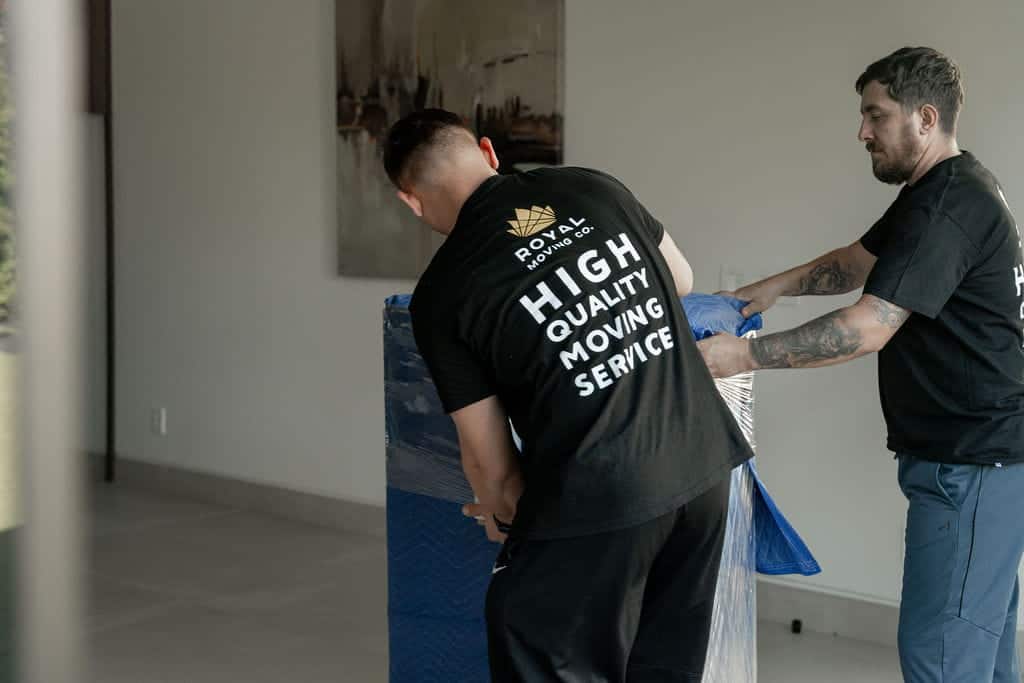 Two movers in matching "High Quality Moving Service" shirts wrap furniture in a blue blanket inside a modern, unfurnished room, showcasing their expert moving services and making it easier to settle in Seattle.