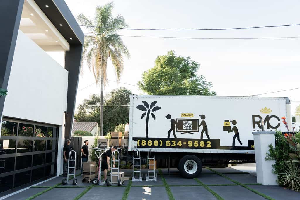 A moving truck is parked in a driveway while movers load boxes using dollies outside a modern house, expertly navigating Seattle to provide top-notch moving services.