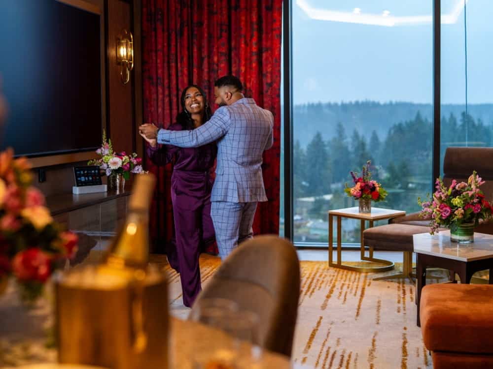 A couple dressed in formal attire dances in a modern, elegantly decorated room with large windows and floral arrangements, creating a romantic atmosphere like nowhere else.